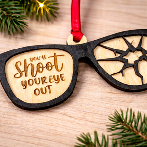 Wooden ornament shaped like sunglasses with text 'you'll shoot your eye out' on a wooden surface with Christmas decorations.