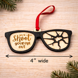 Wooden ornament shaped like sunglasses with text and a red ribbon on a wooden surface.