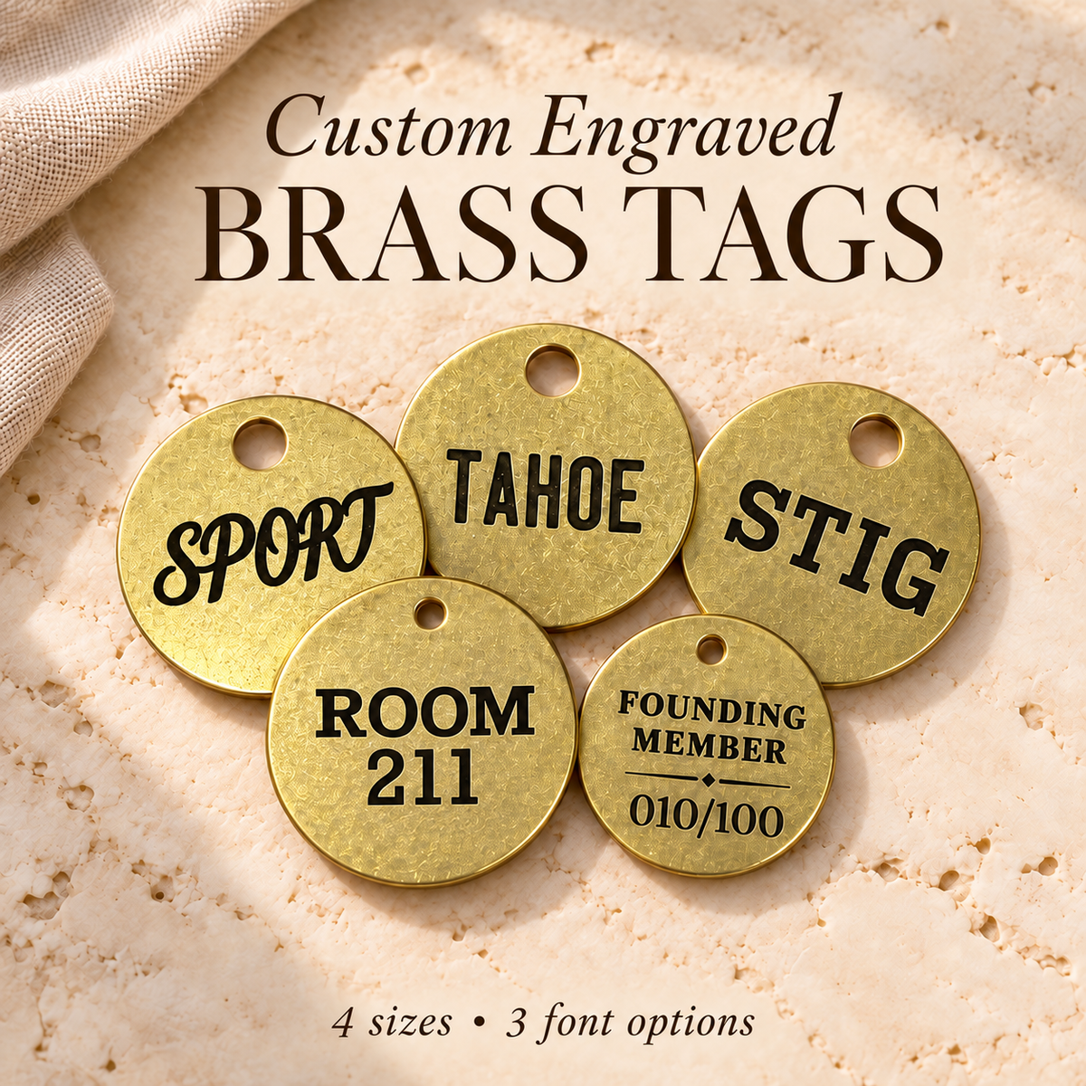Five round brass name tags with pre-drilled holes, shown on a textured beige stone-like surface with fabric in the corner. Large text at the top reads 'Custom Engraved BRASS TAGS,' and smaller text at the bottom says '4 sizes • 3 font options.