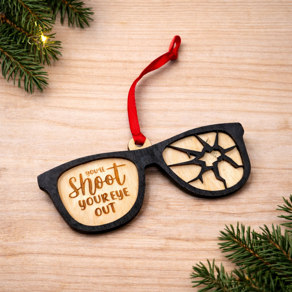 Wooden ornament shaped like sunglasses with a humorous message, hanging on a wooden surface with Christmas decorations.