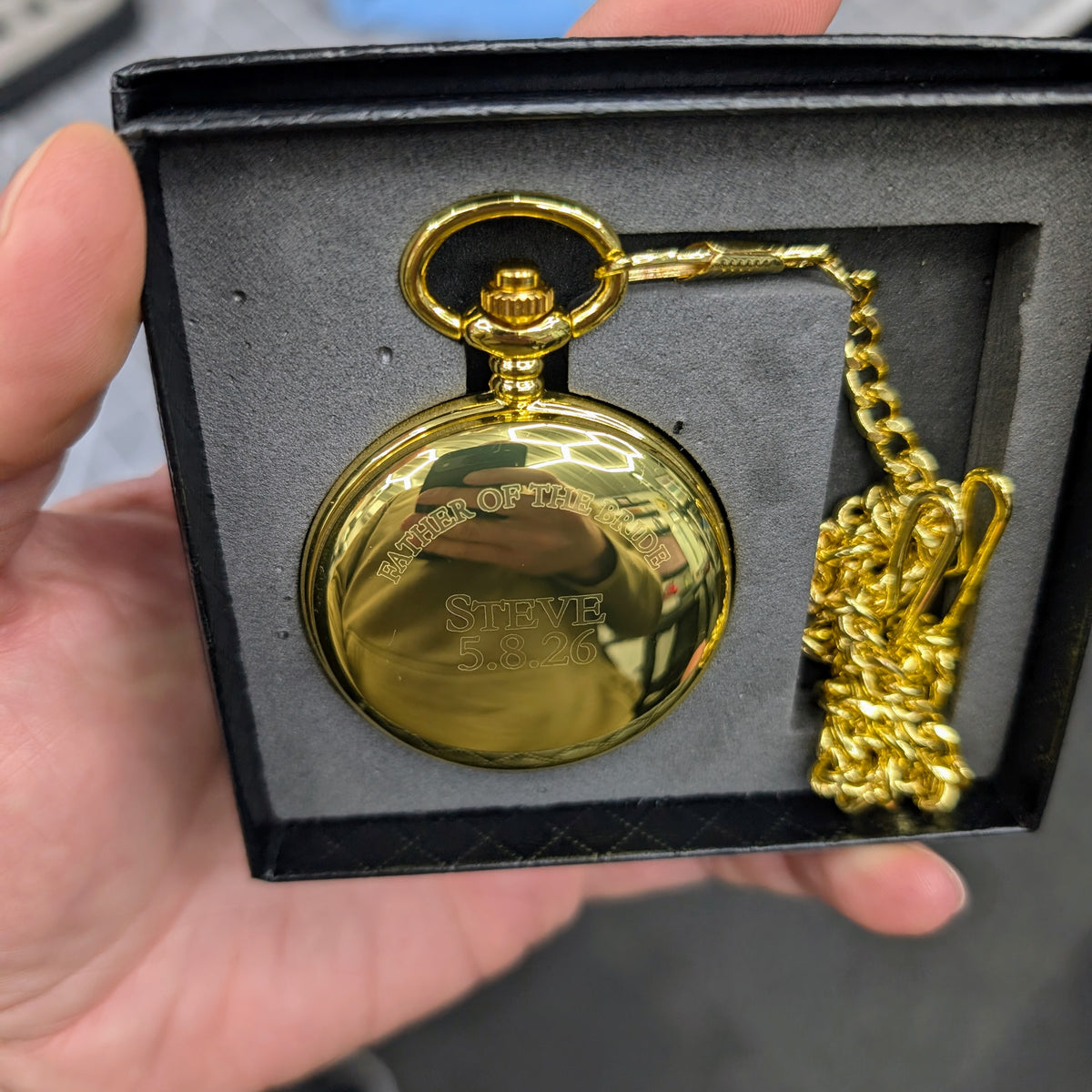 Gold pocket watch with chain in a black box held by a hand.