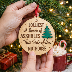 Wooden ornament with humorous text held in front of a Christmas tree and mug.