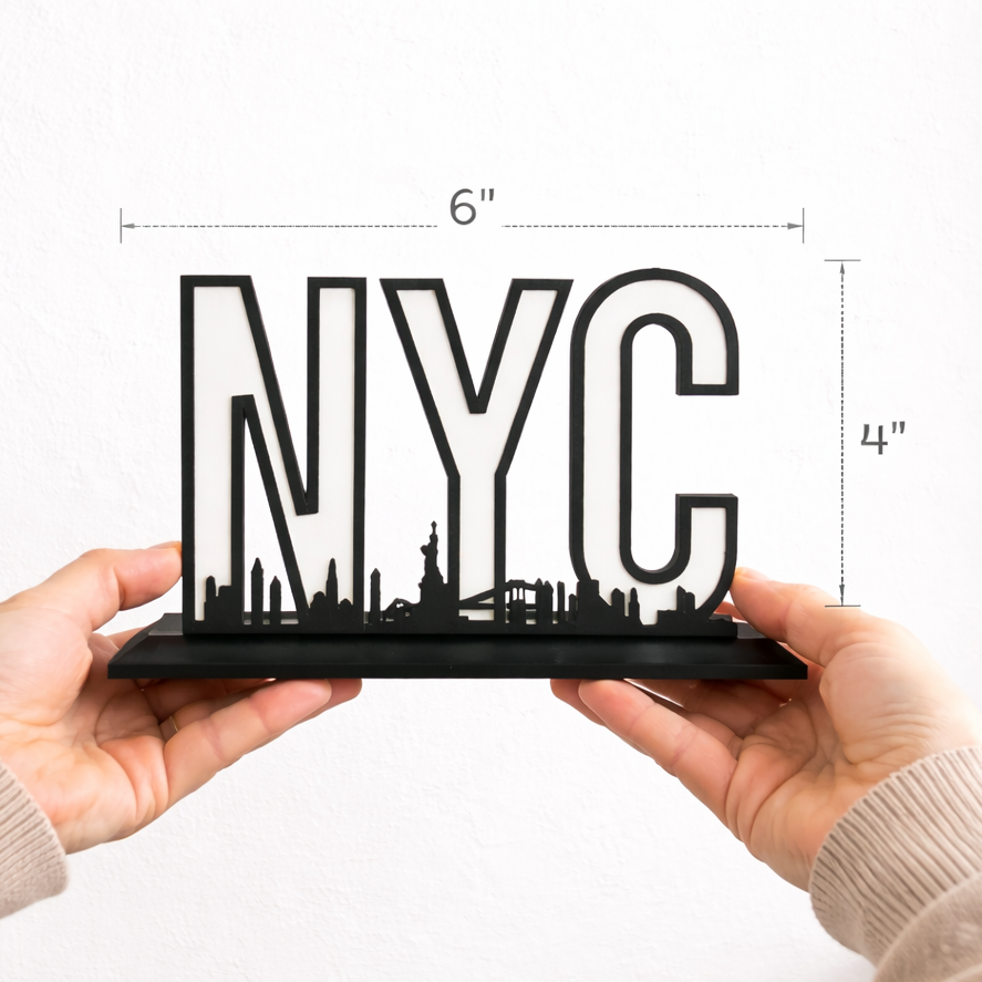 Hands holding a black NYC skyline shelf sign showing true size with measurement lines indicating 6 inches wide by 4 inches tall against a white background.