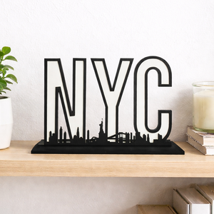 Black NYC skyline shelf sign with bold letter design displayed on a wooden shelf against a white wall, featuring New York City landmarks in a modern minimalist decor setting.